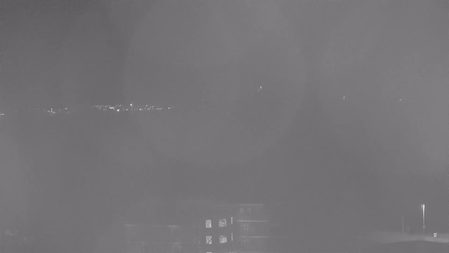 A dimly lit nighttime view captured from a webcam. The foreground shows outlines of structures, possibly buildings or a house, with faint light emanating from a few windows. In the background, a row of distant lights dot the horizon, suggesting a roadway or cityscape. The overall image has a blurred, hazy quality, obscuring some details.