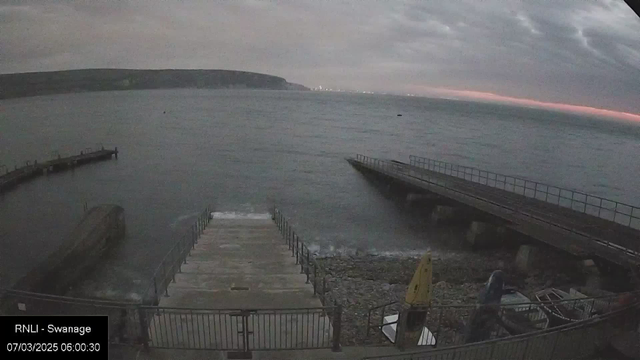 A view of a coastal area at dawn, with overcast skies. In the foreground, there are stone steps leading down to the water, bordered by a low metal railing. To the right, there is a wooden jetty extending into the sea, and a smaller dock adjacent to it. The water appears calm, with gentle waves. Pebbles are visible along the shore, and several boats are moored nearby. In the distance, a faint outline of land can be seen against the lighter horizon, suggesting the approach of sunrise.