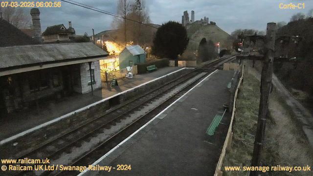 The image shows a quiet railway station during early morning light, with a partially cloudy sky. On the left, there is a stone building with a slanted roof, and a green bench is visible in front of it. Beyond the building, a green shed with lights is partially illuminated. The station platform has several tracks, and a wooden fence runs along the side. In the background, the remnants of a castle can be seen atop a hill, with a few trees and a power pole on the right side of the image. The scene is tranquil, conveying an early hour at the station.