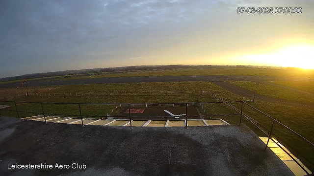 A webcam image capturing a sunrise over a grassy airfield. In the foreground, there is a flat rooftop with a railing. Below, the airfield is visible with a runway extending into the distance. A single small white aircraft is parked on the ground, surrounded by a fenced area. To the left, there is a wind direction indicator near the edge of the field. The sky is partly cloudy, transitioning from dark to bright as the sun rises, casting a warm glow. The date and time in the top right corner indicate March 7, 2025, at 7:00 AM.