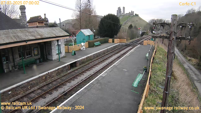 A view of a train station platform with two sets of railway tracks running parallel. The platform features a stone building with a sloped roof, and wooden benches painted green. There is a small green shed to the left and a wooden fence enclosing a grassy area. In the background, hills rise with ruins of a castle at the top, under a cloudy sky. A power pole with wires stands on the right side of the image.