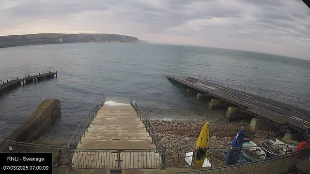 A coastal scene featuring calm water with gentle waves. A concrete ramp leads down to the water, flanked by railings. On the left, there is a small jetty extending into the sea. A rocky shoreline is visible on the right, with several colored kayaks, including yellow, red, and blue, stored along the edge. The background displays distant cliffs under a cloudy sky.