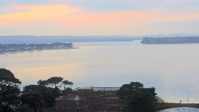 A serene view of a calm body of water at dawn, reflecting shades of orange, pink, and light blue in the sky. In the foreground, there are scattered trees and a low building with several windows. The horizon features a distant shoreline with houses and boats, while gentle hills are visible in the background. The atmosphere is peaceful, suggesting early morning calmness.