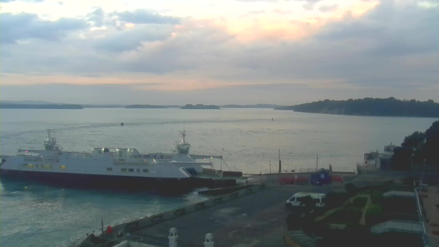 A panoramic view of a harbor at dusk, featuring a large ferry docked at the left side of the image. The water is calm, reflecting the pastel colors of the sky, which is filled with scattered clouds. In the background, several small islands are visible on the horizon, and boats can be seen gently moving across the water. The foreground includes a partially visible dock area with vehicles and some greenery, suggesting a pleasant waterfront atmosphere.