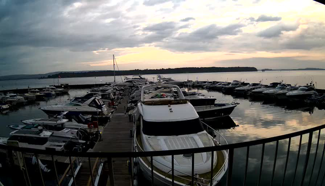 A waterfront scene at a marina, featuring numerous boats and yachts docked along a wooden pier. The foreground shows a large white boat with a cabin. In the background, there are several smaller boats and a calm water surface reflecting the cloudy sky. The sky is overcast, with shades of gray and hints of orange from the setting sun. The landscape includes a distant shoreline with trees and hills.