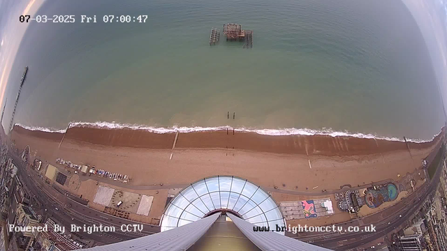 Aerial view of Brighton Beach at sunrise. The scene shows a sandy beach with gentle waves and a structure partially submerged in the water, likely the remains of a pier. The beach is lined with various attractions, including a funfair area. In the foreground, there is a circular observation tower. The image includes a timestamp at the top showing the date and time: March 7, 2025, at 07:00:47. A logo indicates the image is powered by Brighton CCTV.