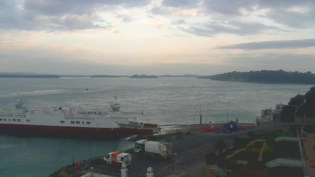 A view of a harbor with a large white and red ferry docked near the shore. In the foreground, there are vehicles parked on a roadway. The background features a calm sea with gentle ripples, and several small islands visible in the distance under a cloudy sky. The colors are muted, suggesting early morning or late afternoon light.