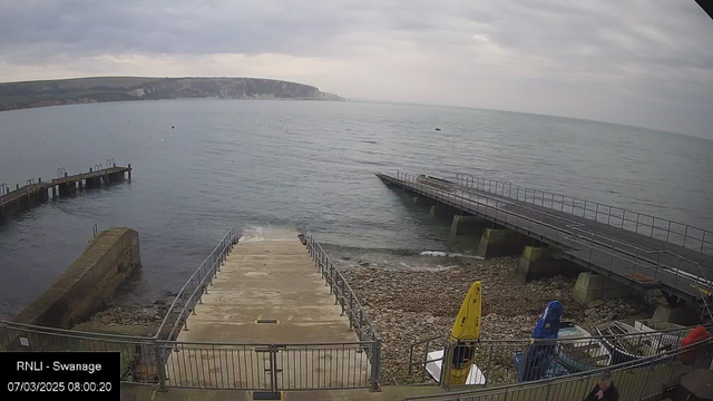 A view of a calm sea with a rocky shoreline and two jetties extending into the water. The sky is overcast with soft, gray clouds. The left jetty is shorter and made of wooden beams, while the right one appears to be longer and made of sturdy materials. There is a set of concrete steps leading down to the water between the two jetties. A yellow kayak is resting on the shore, and several small boats are visible on the water. In the foreground, there are scattered pebbles along the beach area and a figure dressed in dark clothing is seen on the right side near the jetties.