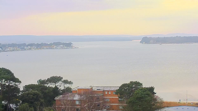 A tranquil waterfront scene showing a wide, calm body of water reflecting soft pastel colors of dawn or dusk. In the foreground, there are trees and a brown building with a flat roof. Across the water, a small collection of boats and a shoreline lined with trees and structures can be seen, leading towards distant hills under a pale sky.