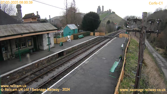 A view of Corfe Castle railway station on a cloudy day. The station features a stone building with a wooden roof, several green benches, and a wooden fence surrounding the area. In the background, Corfe Castle sits atop a hill with a path leading up to it. Two railway tracks run alongside the platform, and a utility pole stands on the right side of the image. There are minimal people present, creating a quiet atmosphere.