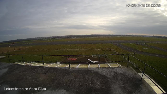 A view from a webcam at Leicester Aero Club shows a large grassy airfield under a cloudy sky. In the foreground, there is a railing and an asphalt path leading down from a building. To the left, a small white aircraft is positioned on the ground. In the background, a wide expanse of green grass is visible, interspersed with a few fences and pathways. The overall atmosphere is calm, with no visible activity on the airfield.