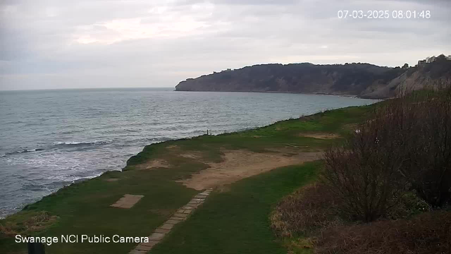 A coastal scene showing a grassy area leading up to a rocky shore. The ocean is visible with gentle waves, and a cloudy sky is overhead. In the distance, a cliff rises, and the landscape appears tranquil with patches of green grass and light brown earth. The image also includes a timestamp in the top right corner.