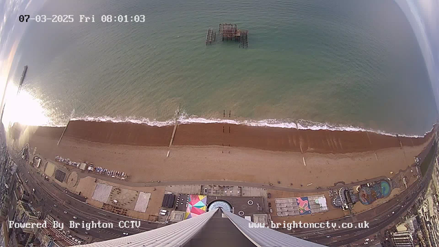 View from a high vantage point overlooking a beach and the sea. The sandy beach stretches horizontally across the image, with gentle waves lapping at the shore. A portion of the view includes a old wooden pier extending into the water. The ocean is calm, reflecting sunlight above. To the right, a variety of beach attractions and structures, including rides and colorful canvases, are visible along the shore, while a road runs parallel to the beach below. The sky is partly cloudy, creating a serene atmosphere. The current date and time are displayed at the top.