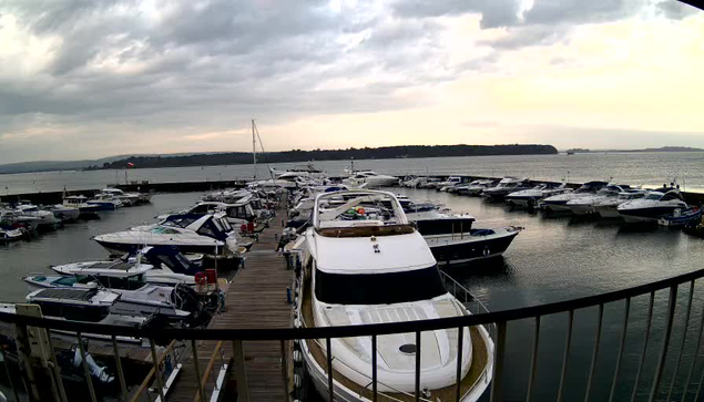 A marina at dusk, featuring numerous boats docked in calm waters. The sky is overcast with gray clouds, and a hint of sunset colors can be seen on the horizon. In the foreground, a white yacht with a dark stripe is prominent, surrounded by various other boats in a mix of colors. A wooden walkway leads from the camera viewpoint to the marina. The atmosphere is tranquil, with gentle waves reflecting the fading light.