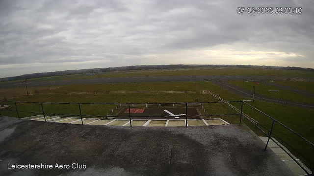 A view from a webcam located at Leicestershire Aero Club, showing a cloudy sky and a large grassy area with a runway in the background. There are some low wooden fences marking the area. The foreground contains a railing and a portion of the building roof. The date and time are displayed in the top right corner of the image.