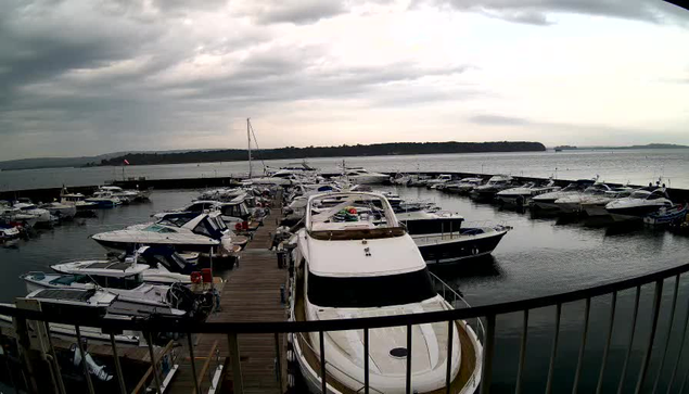 A marina filled with various boats and yachts docked. The scene has a cloudy sky overhead, reflecting a slightly overcast atmosphere. In the background, there is an expanse of calm water, and a distant landmass can be seen on the horizon. The marina is bordered by a wooden walkway, and some boats are moored closely together, while others are more spaced out. The image conveys a serene, nautical environment.