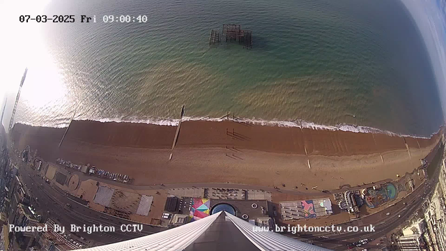 Aerial view of Brighton beach, showing a stretch of brown sand meeting the blue-green water of the sea. There is a ruined pier partially submerged in the water. Along the beach, there are people walking, and amusement park attractions are visible along the promenade, with colorful patches and structures. The image captures a sunny morning atmosphere with clear skies. A timestamp in the corner indicates the date and time.