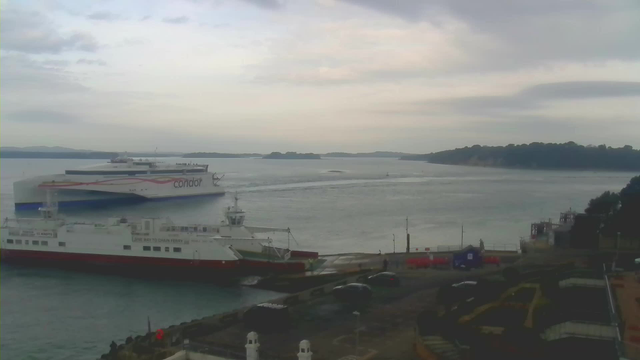 A cloudy sky is visible above a calm body of water. Two ferries are docked; one larger ferry labeled "condor" is departing while a smaller white ferry is moored. The scene includes a shoreline with trees in the background and a few vehicles parked near the water. There are also signs indicating marine traffic rules nearby, and the overall atmosphere appears tranquil and scenic.