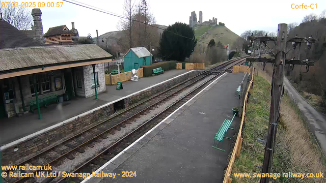 A railway station platform is visible with a single track running alongside it. There are several green benches placed along the platform and a green kiosk in the background. In the distance, a hilly landscape can be seen with ruins of a castle on top. The sky appears overcast, and there are trees lining the area. A wooden fence encloses a section to one side of the platform. A power pole stands on the right, with electrical wires extending overhead.