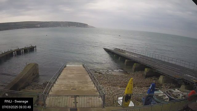 A cloudy seascape showing a shoreline with rocky areas and water. In the foreground, a concrete ramp leads down to the water, bordered by a metal railing. To the right, there are several colorful kayaks resting on the ground. In the background, a pier extends into the water, with a view of steep cliffs in the distance along the coastline. The scene appears tranquil, with gentle waves lapping at the shore.