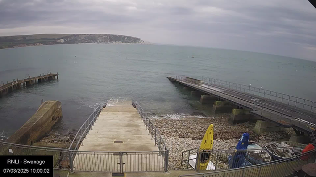 A view of the coastline at Swanage, showing a cloudy sky over the sea. In the foreground, there is a concrete ramp leading down to the water, lined with a metal railing. To the right, a wooden pier extends into the water, and to the left, a structure made of stone is visible. Several boats, including a yellow kayak, are parked on the shore. The water appears calm, with a few buoys floating in the distance.