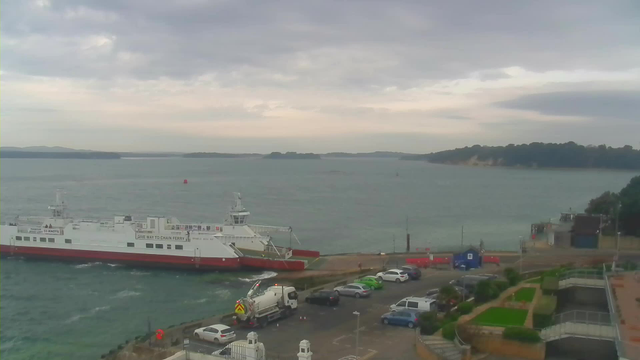 A ferry boat is docked at a pier in a harbor, with water visible around it. The sky is cloudy, and the landscape features distant hills visible across the water. A variety of parked cars can be seen on the waterfront, with some green shrubbery and paved areas nearby.