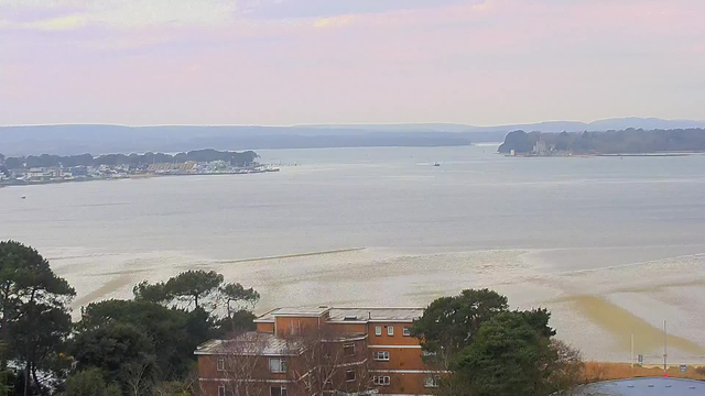 A scenic view of a calm body of water, with distant hills and a cloudy sky. In the foreground, there are trees and a building with a reddish-brown exterior. On the water, small boats can be seen, while the shoreline features a marina with various boats docked. The horizon is lined with trees and hills, creating a tranquil atmosphere.