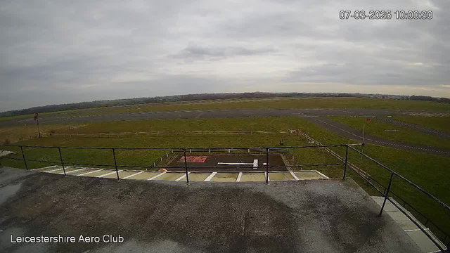 A view from a webcam overlooking a grassy airfield on a cloudy day. In the foreground, there is a low fence along the edge of a building. The airfield features a paved runway that is mainly empty but has some markings and tire tracks visible. The sky is overcast, and the landscape in the distance includes patches of trees and open fields. A small red sign is visible by the runway.