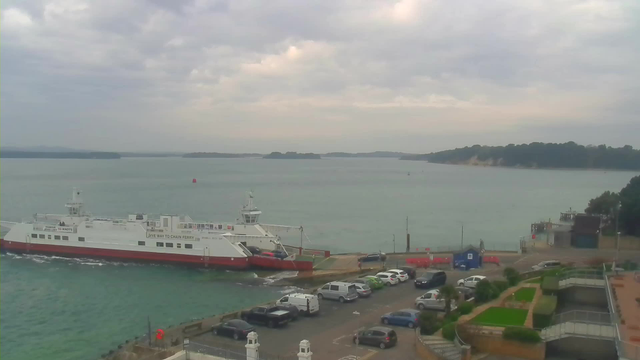 A ferry is docked at a terminal, with vehicles lined up on a parking lot adjacent to the water. The scene features a calm body of water with a distant shoreline visible in the background. The sky is overcast with gray clouds, and there are green and white vehicles parked nearby. Some buildings and a grassy area are seen in the foreground.