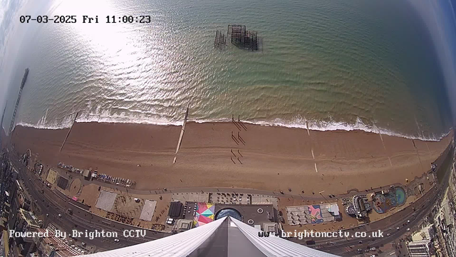 Aerial view of a sandy beach with the ocean waves gently rolling in. In the background, there is a distant pier structure and debris partially submerged in the water. The beach is mostly vacant, with a few people walking along the shore. Alongside the beach, there are colorful attractions and buildings, including a circular design and structures that suggest a seaside amusement area. The sky is partly cloudy with sunlight reflecting off the water, creating a shimmering effect on the ocean's surface. The image is timestamped, indicating the date and time at the top.