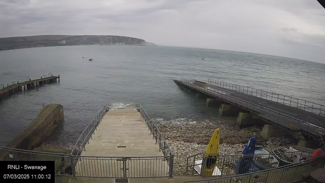 A view of a coastal area featuring a calm sea under a cloudy sky. In the foreground, there is a concrete ramp leading down to the water with a railing. To the left, a wooden pier extends into the sea, and there are a few small boats visible nearby. On the right side, kayaks in yellow, blue, and red are secured to the dock. The backdrop includes grassy hills and cliffs, partially obscured by clouds. The overall scene has a tranquil and serene atmosphere.