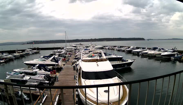 A marina scene featuring numerous boats and yachts docked along a wooden pier. The water is calm, reflecting the cloudy sky above. In the background, there are hills and the shoreline, with a few trees visible. The overall atmosphere is tranquil, with the boats arranged in a neat pattern.