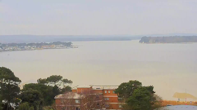 A wide view of a calm body of water, with faintly visible shorelines on both sides. In the foreground, there are some trees and a brown building with multiple windows. The sky is overcast and the atmosphere is hazy, reducing visibility of distant land features. On the left side, some structures and boats are partially visible along the shoreline. The water appears to be a muted color, likely reflecting the gray sky.