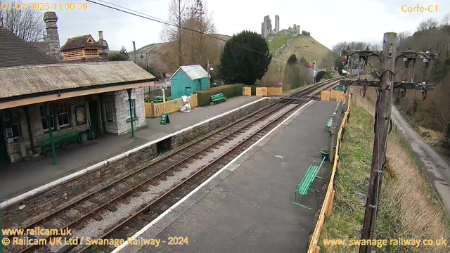 A view of a railway station with a stone building featuring a sloped roof and several windows. Green benches are placed along the platform, and a sign indicates "WAY OUT." In the background, a green hillside rises, with castle ruins visible at the top. A wooden fence surrounds part of the area, and a telephone pole with wires stands to the right. The scene is set on a cloudy day, suggesting a typical overcast sky.