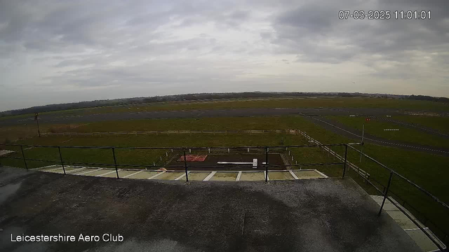 A cloudy sky with patches of grey covers the scene. In the foreground, there is a flat rooftop with a railing, showing a wide and empty grassy area below. The landscape includes a runway, which is mostly bare, and a few scattered markers or signs along its edge. The background features a gentle slope of land, leading to a distant horizon where low hills are visible. The time and date are displayed in the top right corner, indicating 11:01 AM on March 7, 2025.