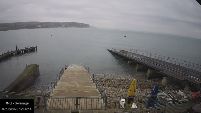 A view of a calm sea under a cloudy sky, with a rocky shoreline on the left. There is a concrete ramp leading down to the water, flanked by railings. To the right, a wooden pier extends into the water, and a second, narrower pier is visible further in the distance. Two kayaks in bright yellow and blue are stored on the shore beside the ramp. A person is standing on the pier, looking out at the water.