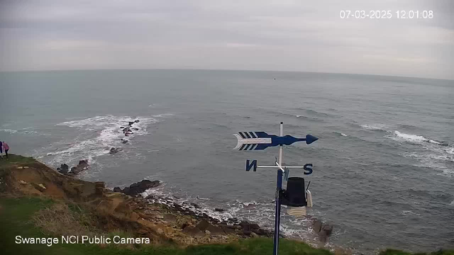 A view of the coastline at Swanage, featuring the sea with gentle waves and patches of rocky land emerging from the water. In the foreground, there is a grassy area with the hint of a path leading towards the edge. A weather vane with blue arrows is positioned on the right side, showing north and south directions. The sky is overcast, with soft gray clouds, creating a tranquil atmosphere. The date and time displayed in the corner indicate it is March 7, 2025, at 12:01 PM.