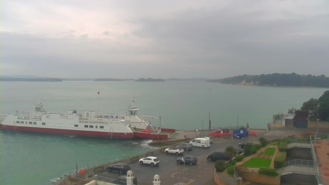 A ferry with a white body and red lower section is docked at a harbor, partially submerged in turquoise water. The background features a slightly cloudy sky and green landscape in the distance with hills. In the foreground, there is a parking lot with several cars, including white and black vehicles, alongside a walkway and landscaped areas with grass and shrubs.