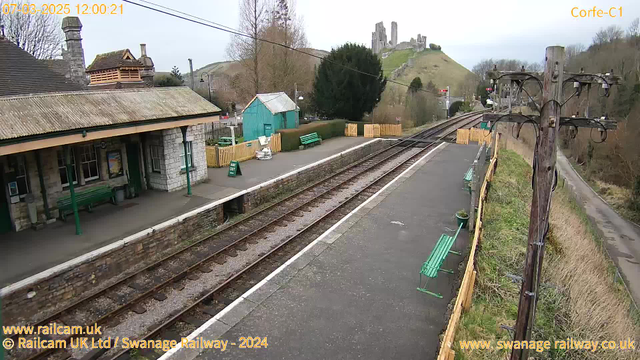 A railway station platform is visible with a stone building featuring a peaked roof on the left. There are several green benches along the platform. In the background, there is a hill with ancient ruins at the top, and a small blue shed is situated on the platform. The railway tracks run parallel to the platform, and there are wooden fences and signs in the area. The sky is overcast, and trees are present in the scene, along with a power pole on the right.