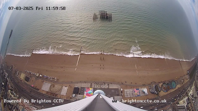 Aerial view of a beach with golden sand and gentle waves lapping at the shore. In the background, a pier extends into the water, with some structural remnants visible. The beach is populated with small groups of people walking, and various colorful beach structures can be seen along the shoreline. The image displays a clear sky with scattered clouds, and the timestamp at the top indicates the date and time. The lower part of the image features an amusement park area with attractions, including a pool and game stalls.