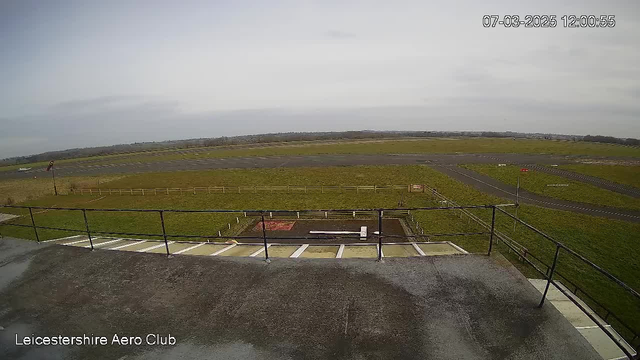 A view from a high vantage point at Leicestershire Aero Club. In the foreground, there is a flat rooftop area with a railing. Below, a grassy field extends across the scene, bordered by wooden fencing. There are multiple runways visible, mostly paved, with some areas of grass in between. A few small objects are positioned on the ground near the center. The sky is mostly cloudy with a soft light, indicating daytime.