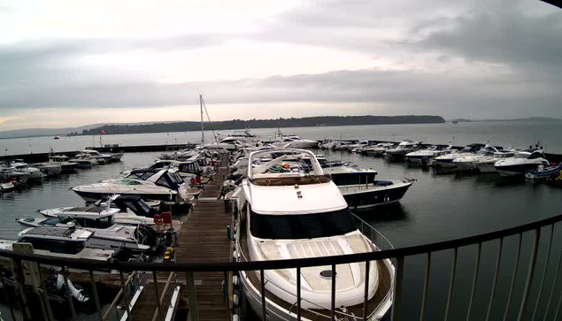 A marina scene featuring multiple boats docked in calm water. The foreground showcases a large white yacht with a sleek design, while smaller boats, some with blue and white stripes, are moored nearby. The background has a cloudy sky, and the water reflects the overcast ambiance. There is a wooden dock visible in front, with several boats lined up along it. The overall atmosphere is tranquil, typical of a harbor setting.