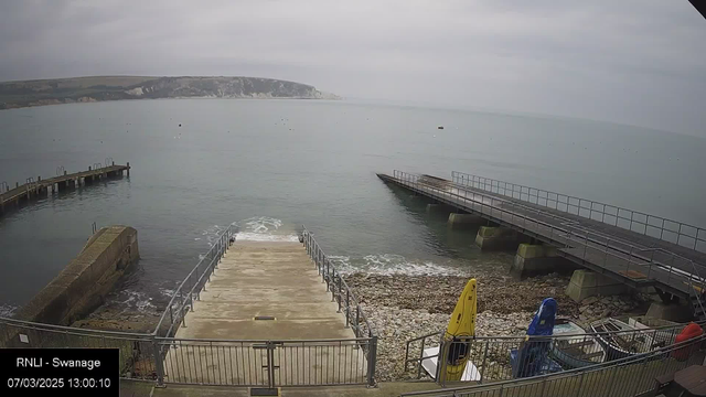 A coastal scene showing a body of water with calm waves, a sandy and rocky shoreline, and a cloudy sky above. In the foreground, there is a concrete ramp leading down to the water, along with a railing. To the right, there are two boat docks extending into the water, one with a flat surface and the other slightly elevated. Several boats and a yellow kayak are visible on the shore. The overall atmosphere is serene and tranquil.