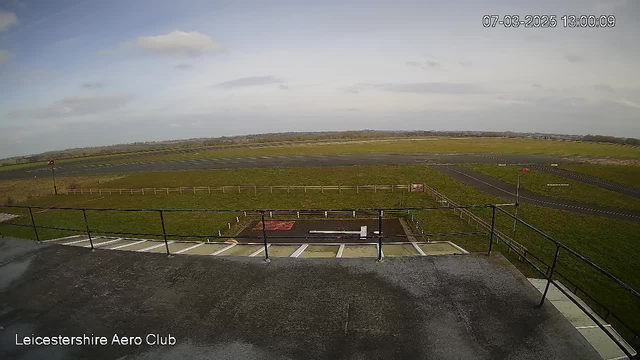 A view from a high vantage point at the Leicestershire Aero Club, showing a wide expanse of grass and asphalt airstrip under a cloudy sky. In the foreground, there is a railing with a rooftop surface below. The airstrip has a few markings, and a hangar is visible in the distance. Farmland stretches out beyond the airstrip, with scattered trees and a light blue horizon. The scene indicates a cool, overcast day.