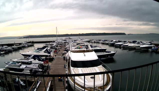 A marina filled with various boats docked neatly in a calm body of water. The scene shows a mix of pleasure craft and larger yachts, with some boats reflecting light on the water’s surface. The sky is overcast, with soft gray clouds, and hints of lighter areas near the horizon, suggesting the sun is either rising or setting. A wooden walkway or dock is visible in the foreground, leading towards the boats. In the distance, a tree-lined shore can be seen on the right.