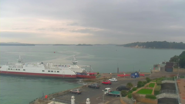 A cloudy sky looms over a harbor scene with calm water. A white ferry with a red lower half is docked at the edge of the pier. Several cars are parked nearby on the asphalt, including a red car and some larger vehicles. In the distance, small islands can be seen, and there's a low shoreline with greenery. A portion of the pier is visible to the right, with a blue building and some red barriers. The atmosphere is serene, with gentle waves lapping against the dock.