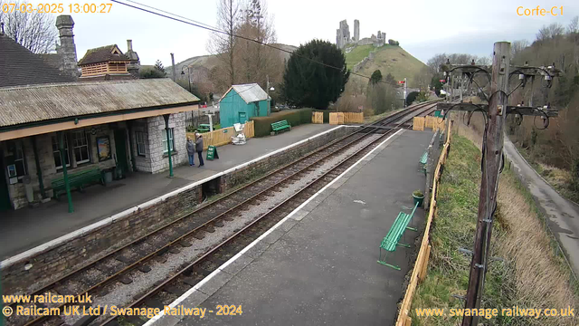 The image shows a railway station platform with a stone building on the left. Two people are standing near a green sign that reads "WAY OUT." There are green benches along the platform and a wooden fence in the background. On the hill in the distance, there is a castle ruin. Power lines run along the platform on the right side of the image. The scene is set during the day with a cloudy sky.