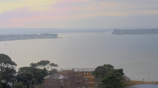 A serene coastal view at dawn. The calm water reflects soft pastel colors of pink and purple from the sky. In the distance, a small marina with boats is visible, and a wooded shoreline borders the left side. In the foreground, there are low trees and a building with a flat roof, appearing partially obscured by vegetation. The scene conveys a peaceful, early morning atmosphere.