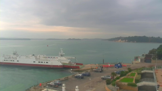 A large white and red ferry is docked at a harbor, with calm waters surrounding it. The sky is overcast with gray clouds, and the horizon shows distant land with trees. In the foreground, there are several parked cars, and a small blue building is visible by the dock. There are some people near the ferry, and the area seems quiet with a green landscaped section on the right side.