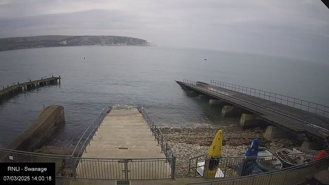 A serene coastal view featuring a set of stairs leading down to the water, alongside a rocky shoreline. In the background, a long wooden pier juts out into the calm sea. Two brightly colored kayaks, one yellow and one blue, are positioned on the pebbly beach. The sky is overcast, with soft gray clouds, and the distant cliffs can be seen on the horizon.